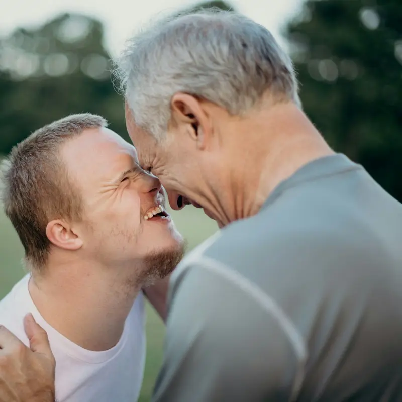 A young man and his father touching foreheads and laughing outdoors